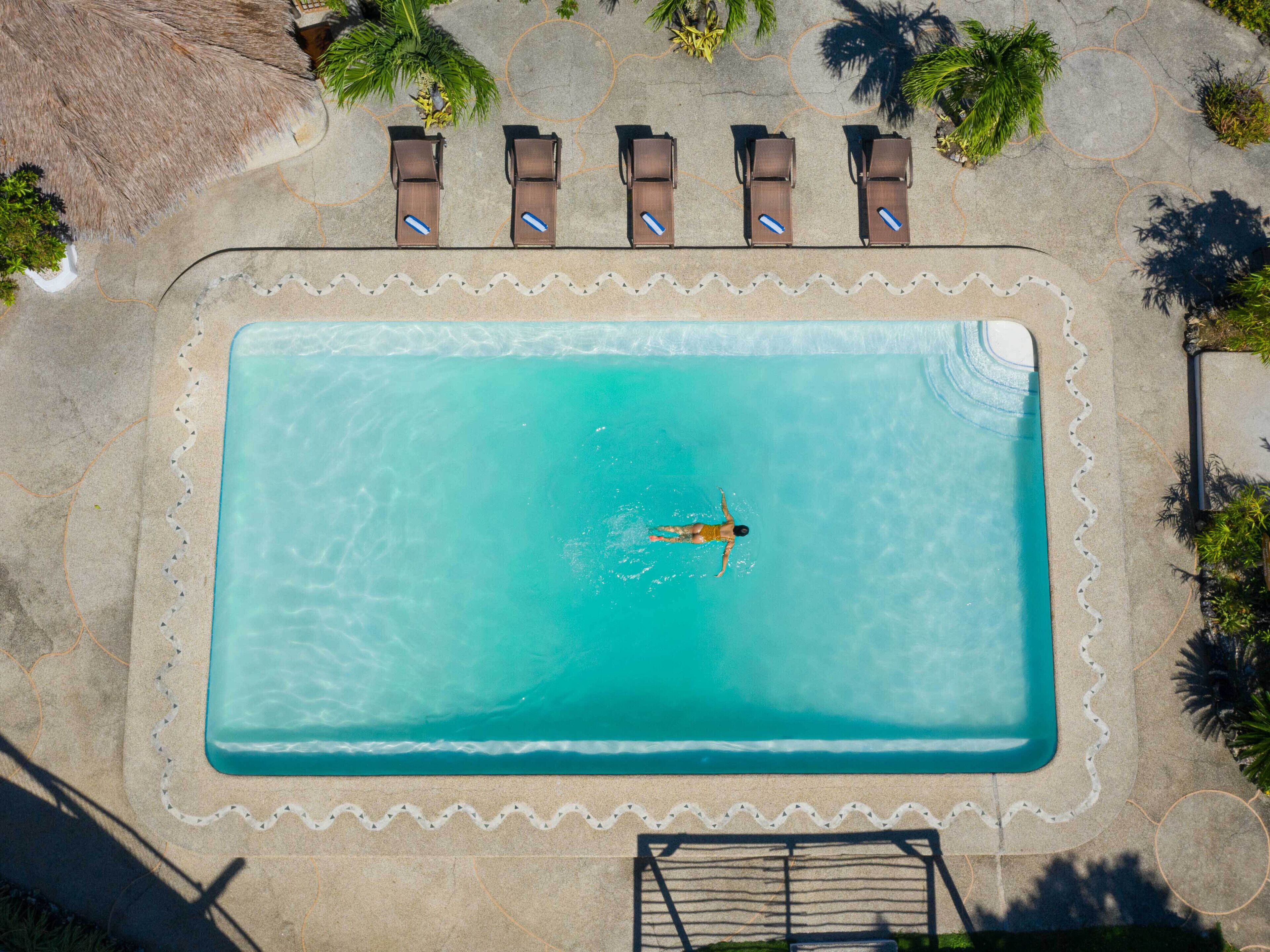 An aerial view of a rectangular swimming pool with a person swimming, surrounded by lounge chairs and palm trees.