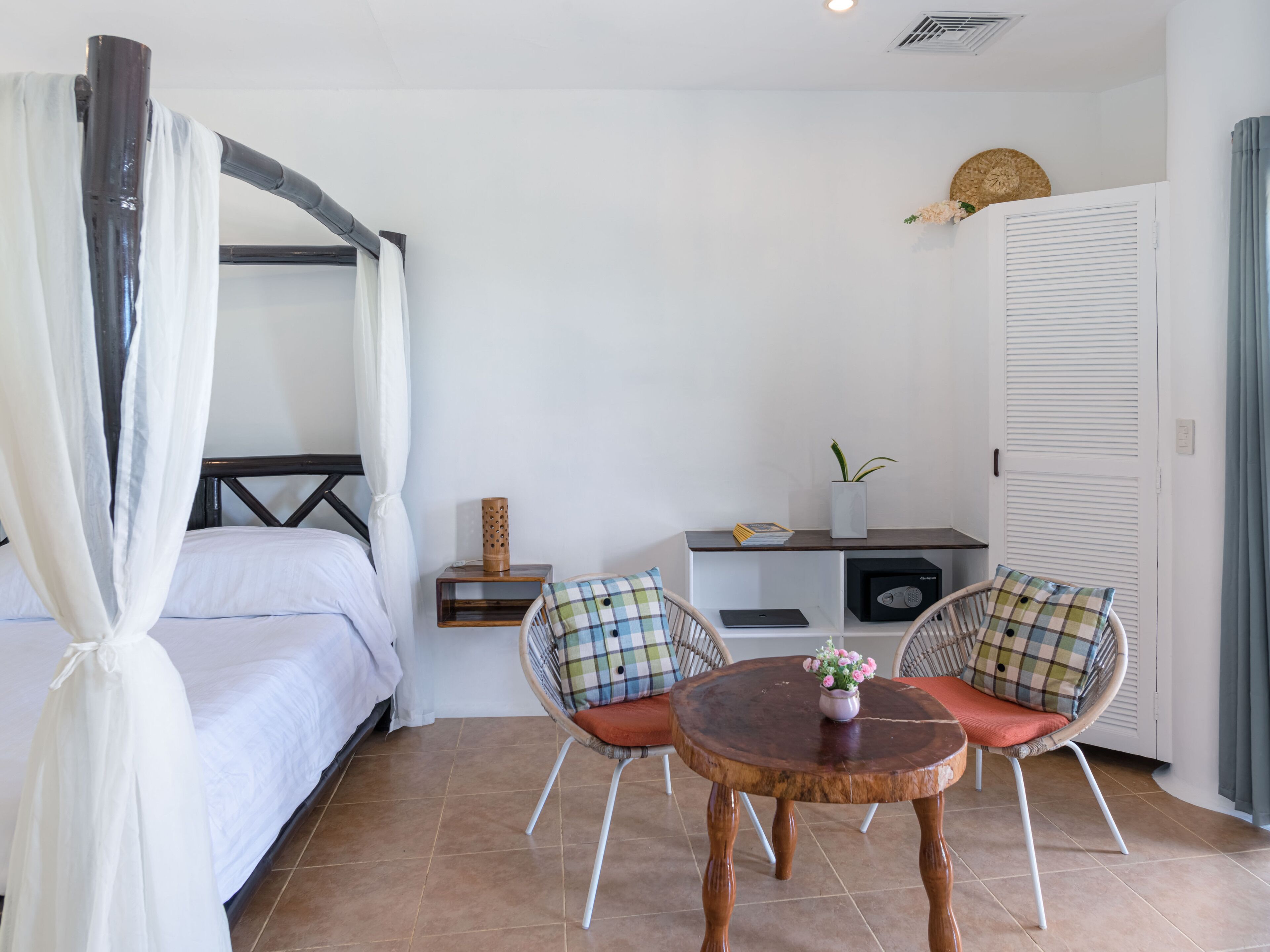 A bedroom with a four-poster bed, a bedside table with a lamp, a small shelf with decorative items, and a chair with a cushion.