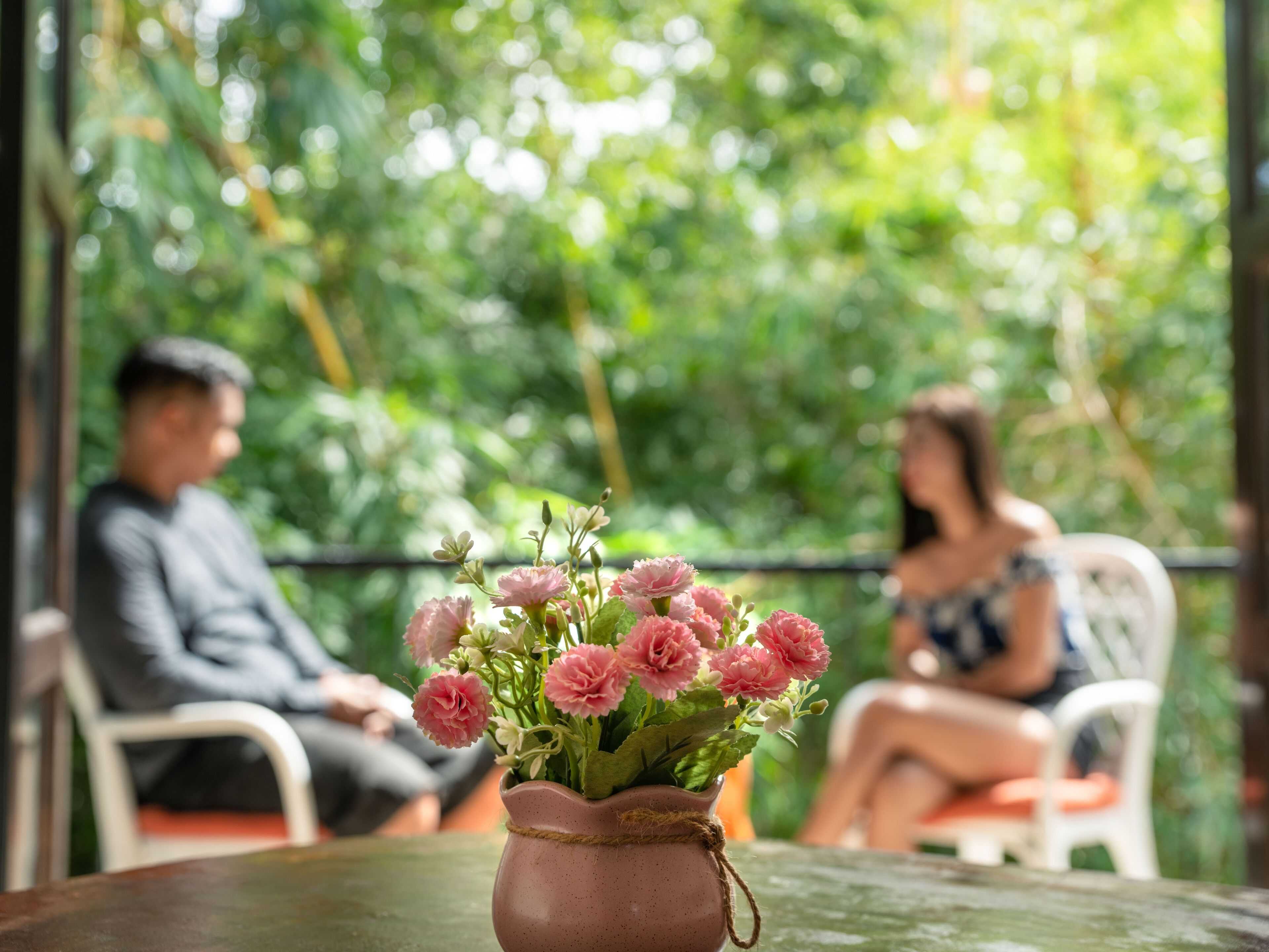 Una mesa con un jarrón de flores rosadas, dos personas sentadas en sillas blancas y un fondo verde exuberante.