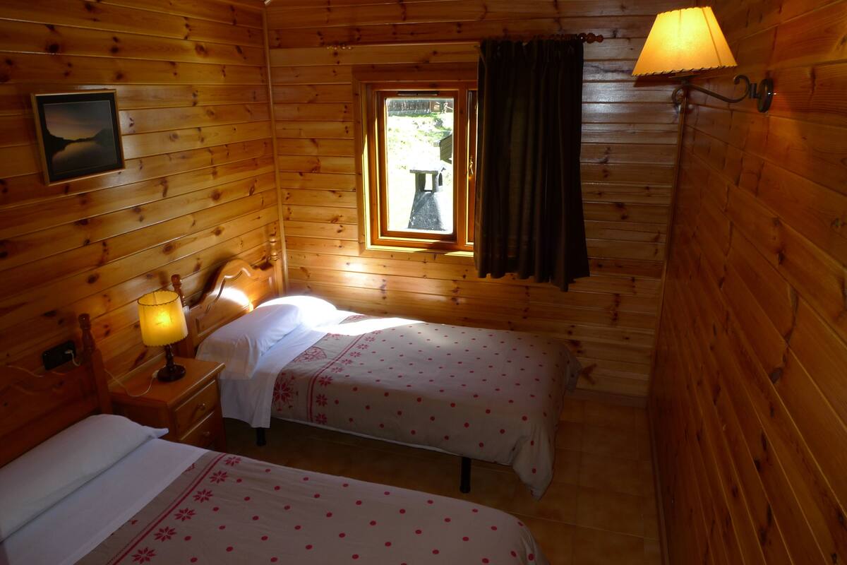 Une chambre de style cabane en bois, avec deux lits, une fenêtre avec des rideaux, une table de chevet avec une lampe et un tableau encadré au mur.