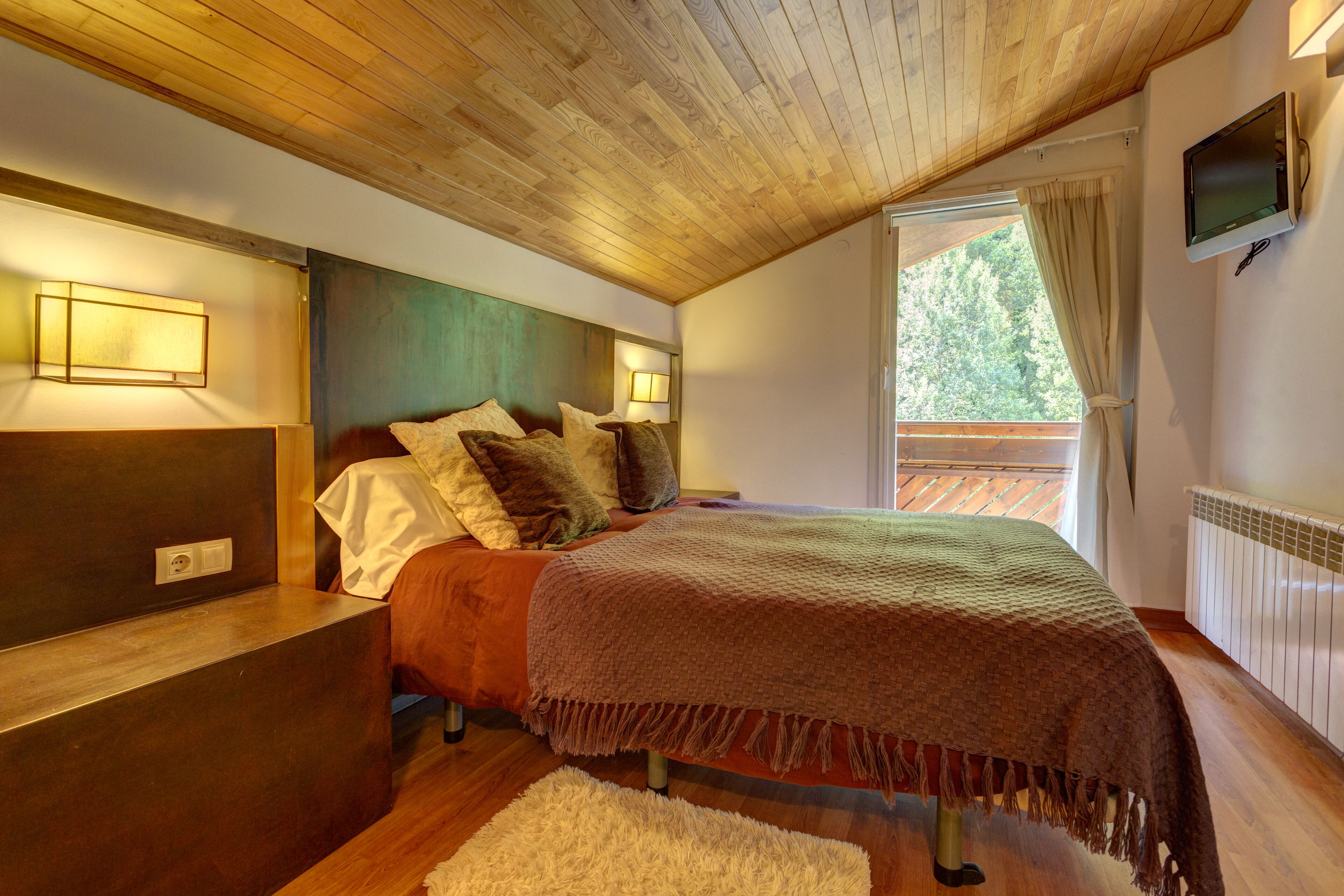 Een slaapkamer met een houten plafond, een bed met een bruine deken, een nachtkastje, een tv aan de muur en een raam met gordijnen.