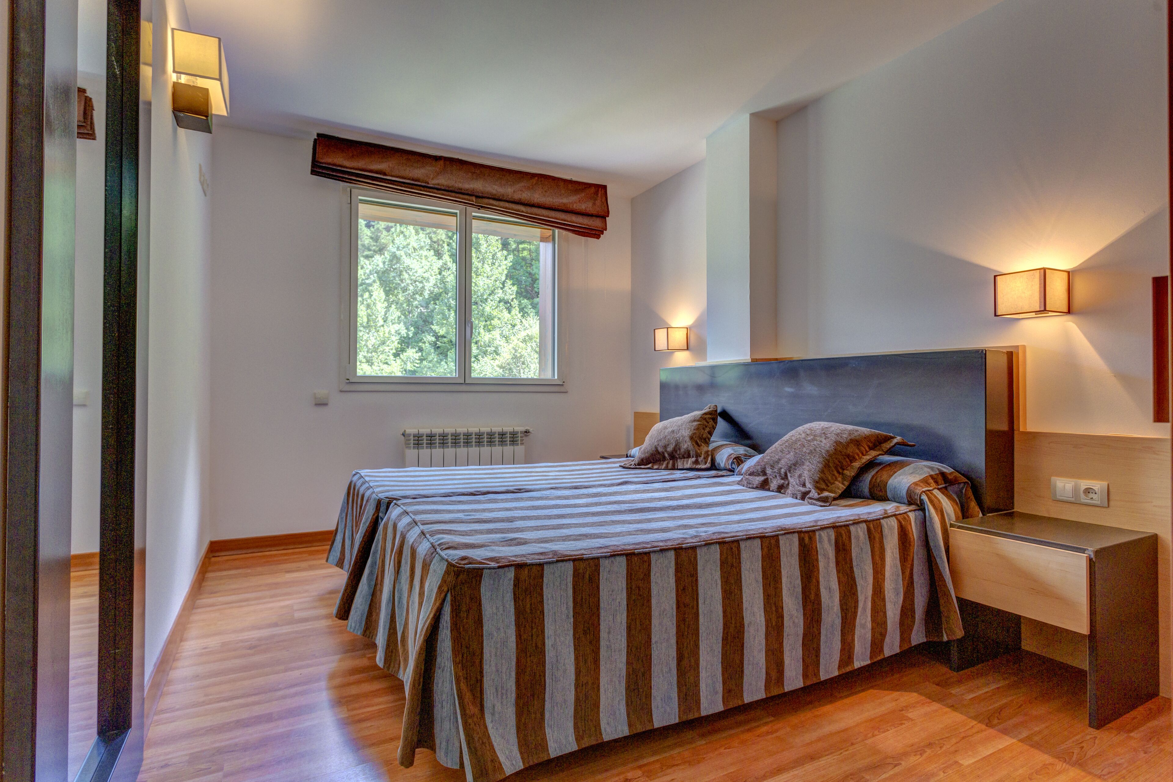 Un dormitorio con una cama doble, dos mesitas de noche, un ventanal con vistas a árboles y lámparas de pared.