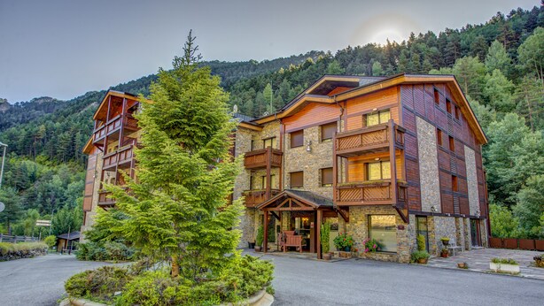 A multi-story lodge with a stone facade and wooden balconies, surrounded by lush greenery and a mountainous backdrop.