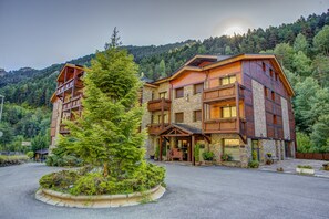 A multi-story lodge with a stone facade and wooden balconies, surrounded by lush greenery and a mountainous backdrop. - Apartamentos y Bungalows Xixerella Park (La Massana)