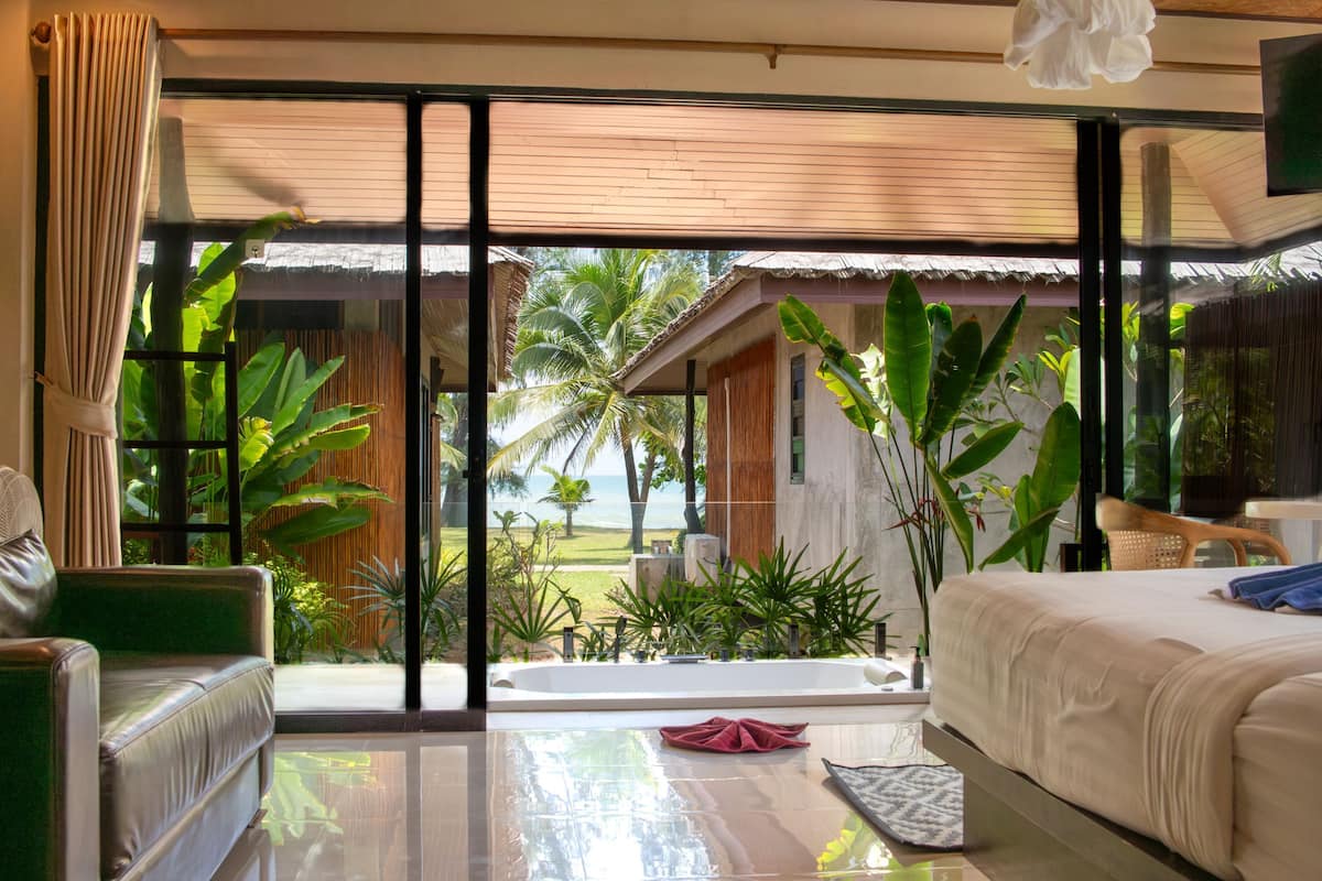 A spacious room with a large sliding glass door leading to a balcony with a view of palm trees and a beach. The room includes a bed, a green sofa, and a small table.