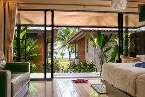 A spacious room with a large sliding glass door leading to a balcony with a view of palm trees and a beach. The room includes a bed, a green sofa, and a small table.