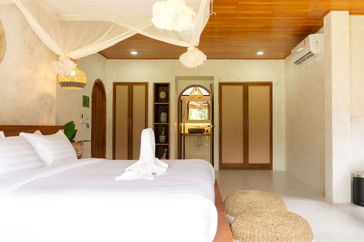 A bedroom with a large bed, a bedside table, a woven pouf, a wooden ceiling, and a window with a view of the outdoors.