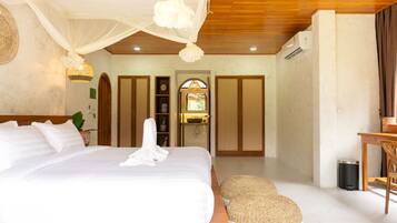A bedroom with a large bed, a bedside table, a woven pouf, a wooden ceiling, and a window with a view of the outdoors.