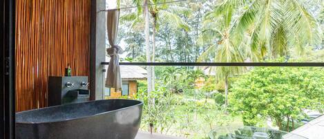 A modern bathroom with a large freestanding bathtub, a glass table, and a view of a lush tropical garden.