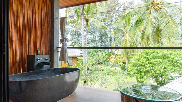 A modern bathroom with a large freestanding bathtub, a glass table, and a view of a lush tropical garden. - Long Beach Chalets (Ko Lanta)