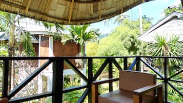 A wooden deck with a thatched roof, surrounded by lush greenery and a view of a traditional house.