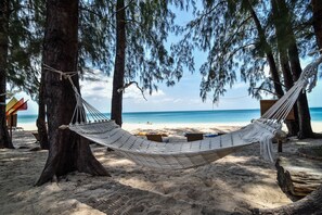 A hammock tied between two trees on a sandy beach with a clear view of the ocean.