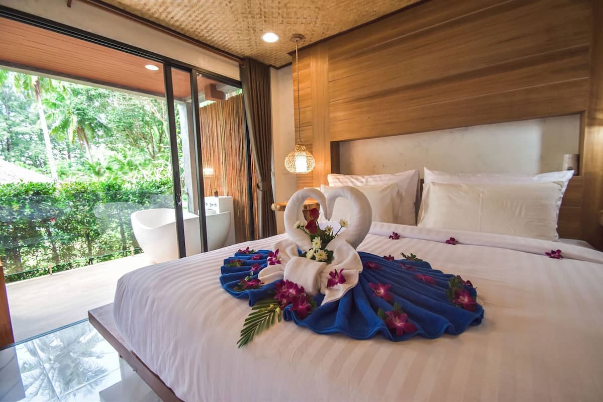 A hotel room with a large bed, a wooden headboard, a glass partition, and a view of greenery outside.