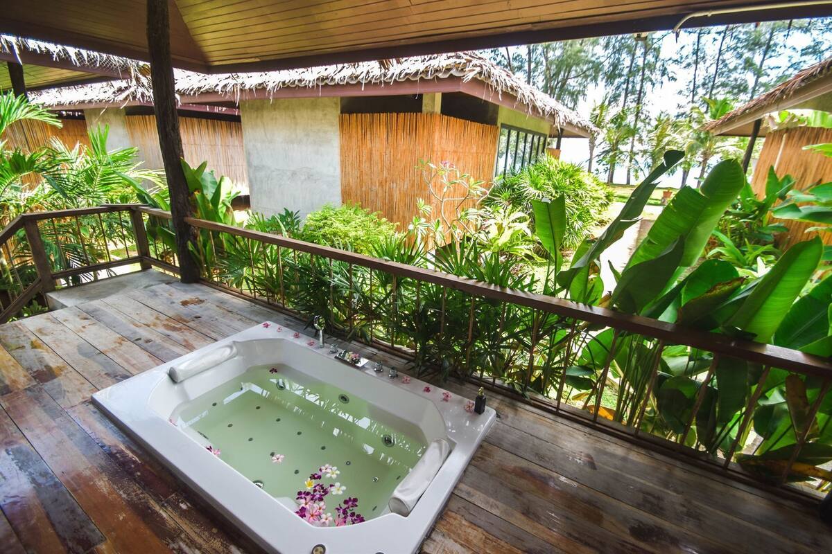 A wooden deck with a hot tub, surrounded by lush tropical plants and a thatched-roof structure.