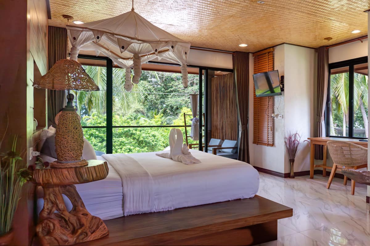 A bedroom with a large bed, a wooden bench, a TV, and a balcony with a view of greenery.