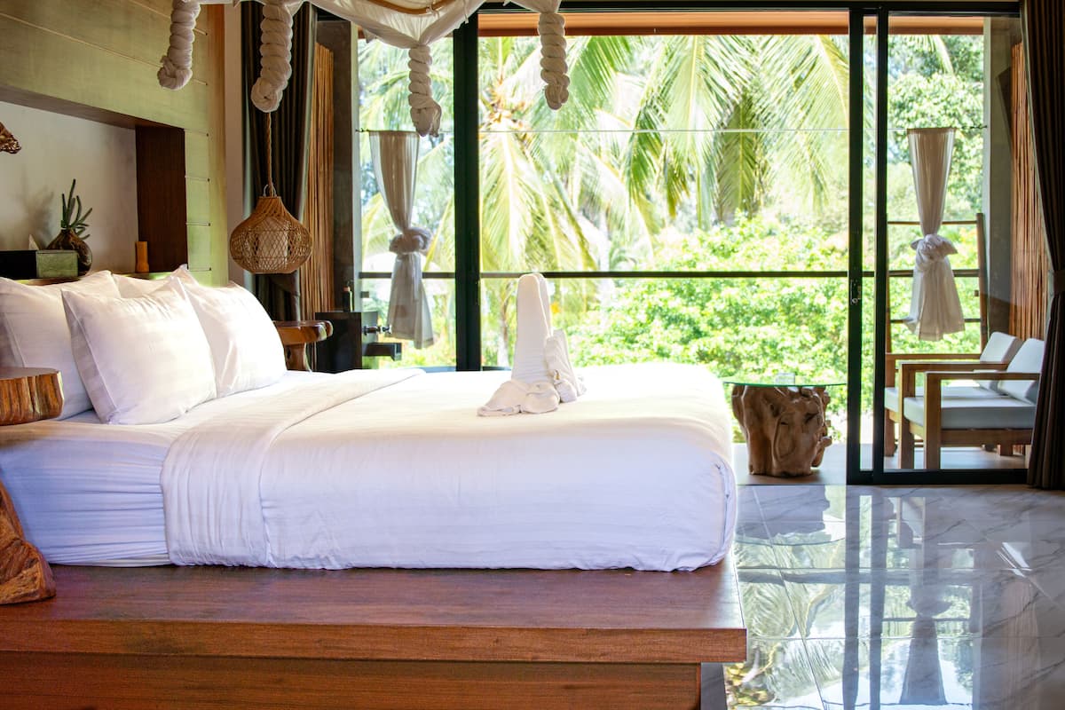 A spacious bedroom with a large bed, a wooden bench, and a view of lush greenery through large windows.