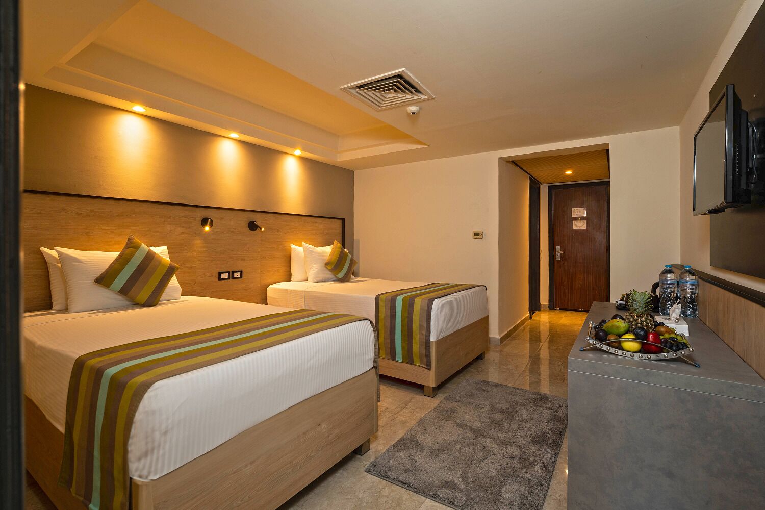 A hotel room with two beds, a desk with fruit, and a TV.