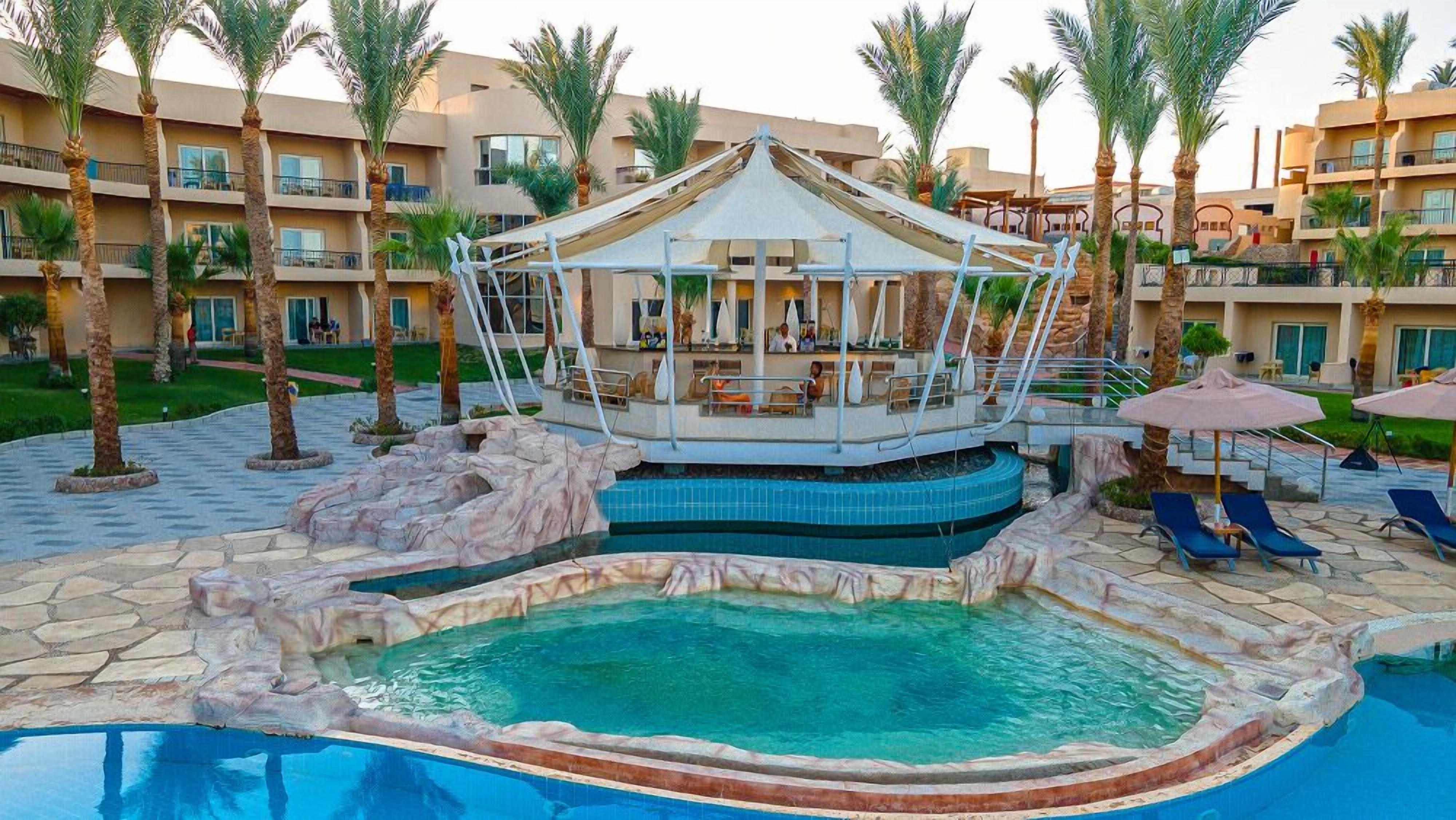 A resort pool area with a central water feature, lounge chairs, and a covered seating area.