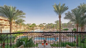 A balcony with two chairs and a table, overlooking a pool area with palm trees and a building complex. - Xperience Kiroseiz Premier (Sharm El Sheikh)