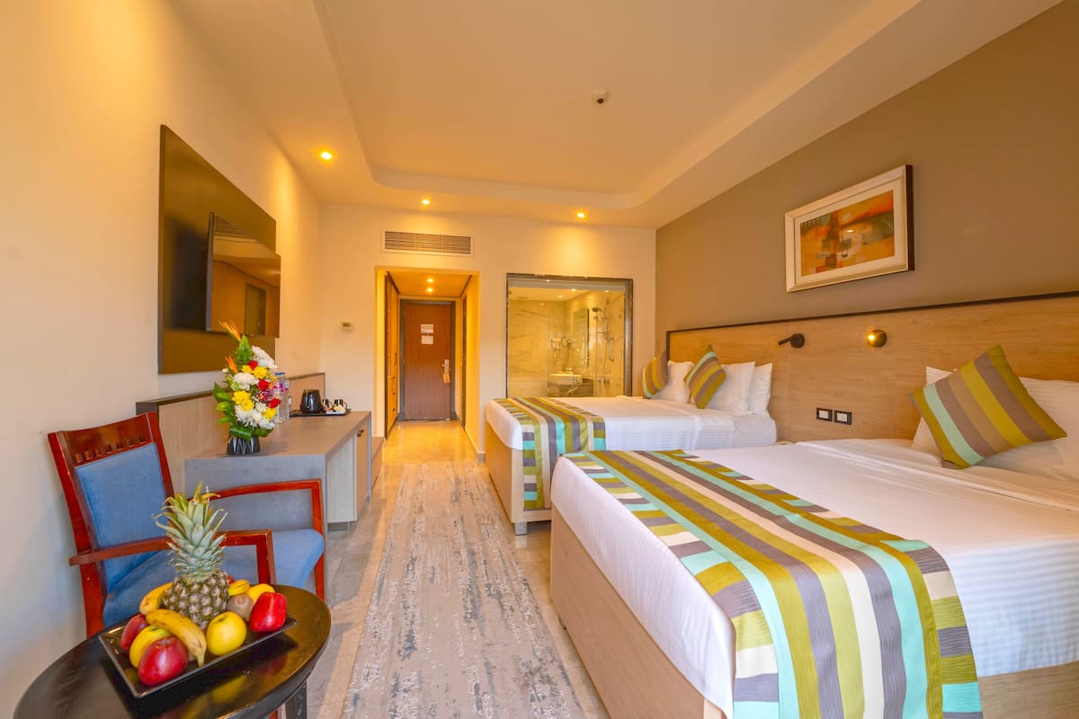 A hotel room with two beds, a desk with a chair, a fruit bowl, and a shower area.