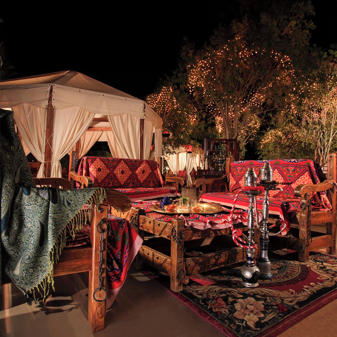 Un espacio al aire libre con carpas, muebles de madera tradicionales, una mesa de comedor con platos y un narguile, y un árbol decorado con luces en el fondo.