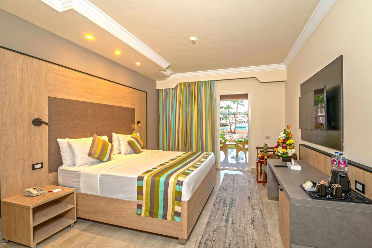 A modern hotel room with a large bed, a bedside table with a phone, a desk with a coffee maker, and a TV on the wall.