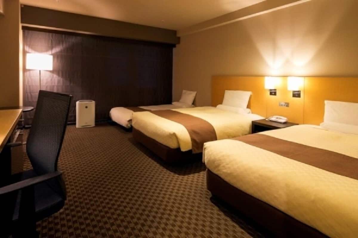 A hotel room with two beds, a desk, and a chair. There are two bedside tables with lamps and a wall-mounted phone.