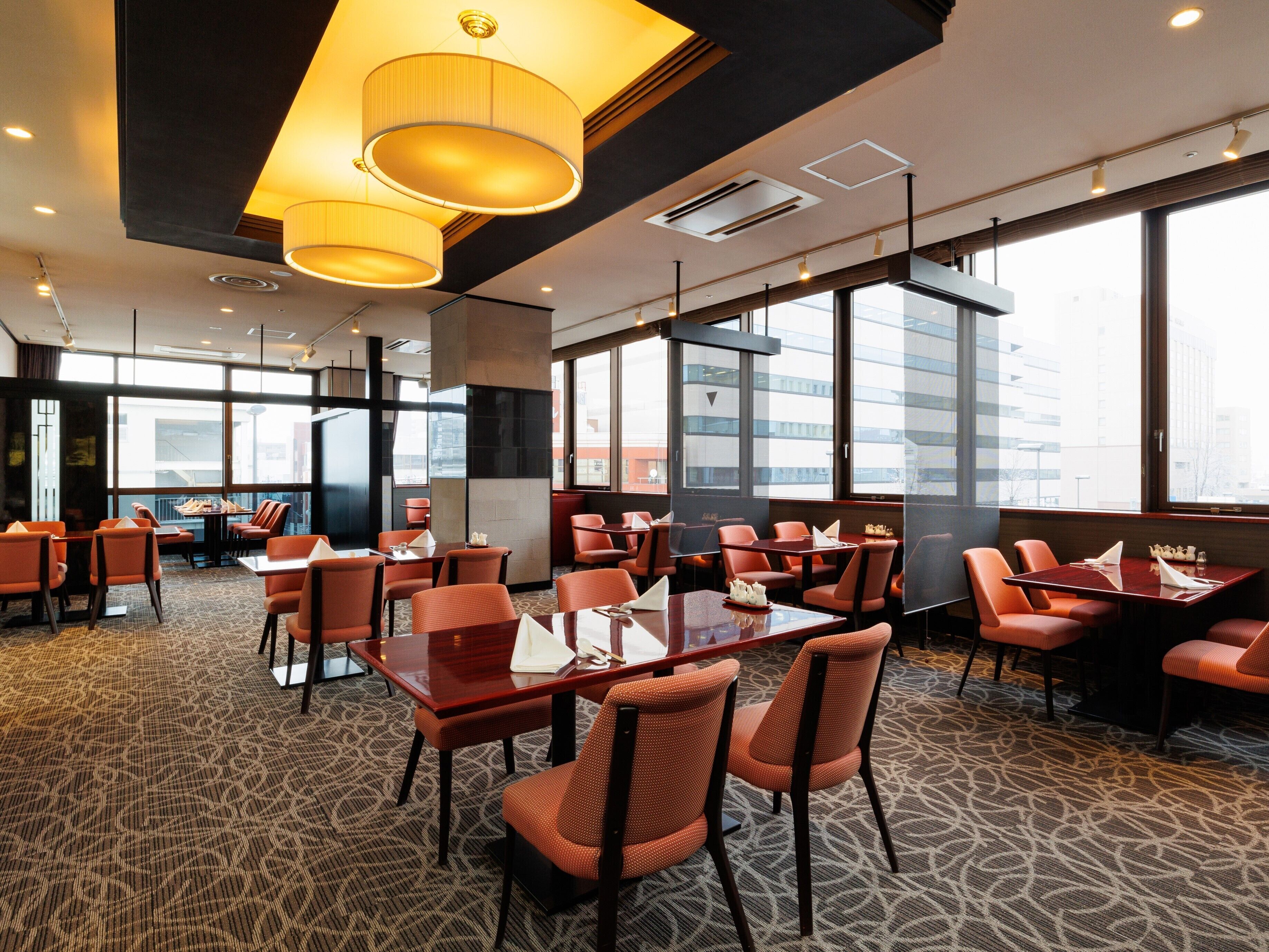 Un comedor de hotel moderno con techos altos, ventanas amplias y sillas tapizadas en naranja.