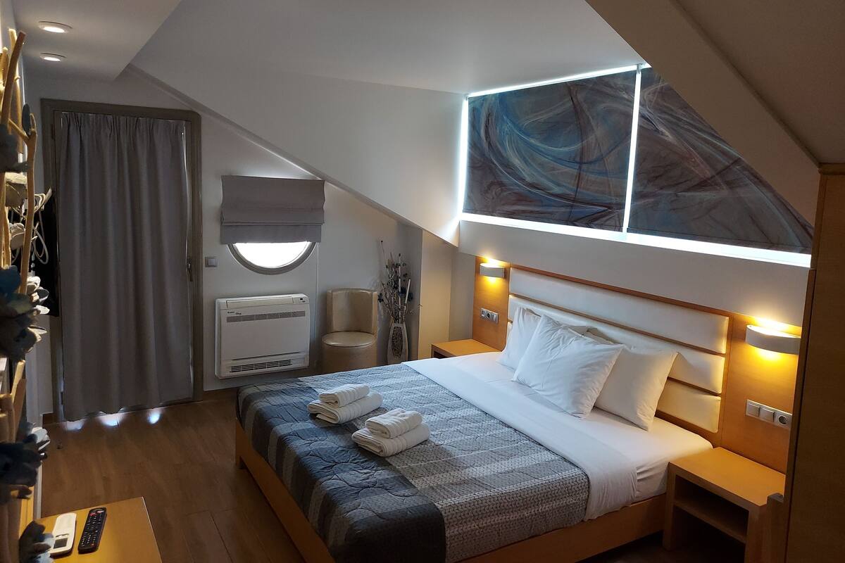 Ein modernes Hotelzimmer mit Bett, Nachttischen, einem Schreibtisch, einem Sessel und einer Klimaanlage. An der Wand hängt ein großes, abstraktes Gemälde.