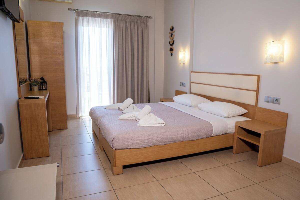 Ein ordentlich eingerichtetes Hotelzimmer mit einem Holzbett, einem Nachttisch, einem kleinen Holzbeistelltisch, einem Spiegel und einem Fenster mit Vorhängen.
