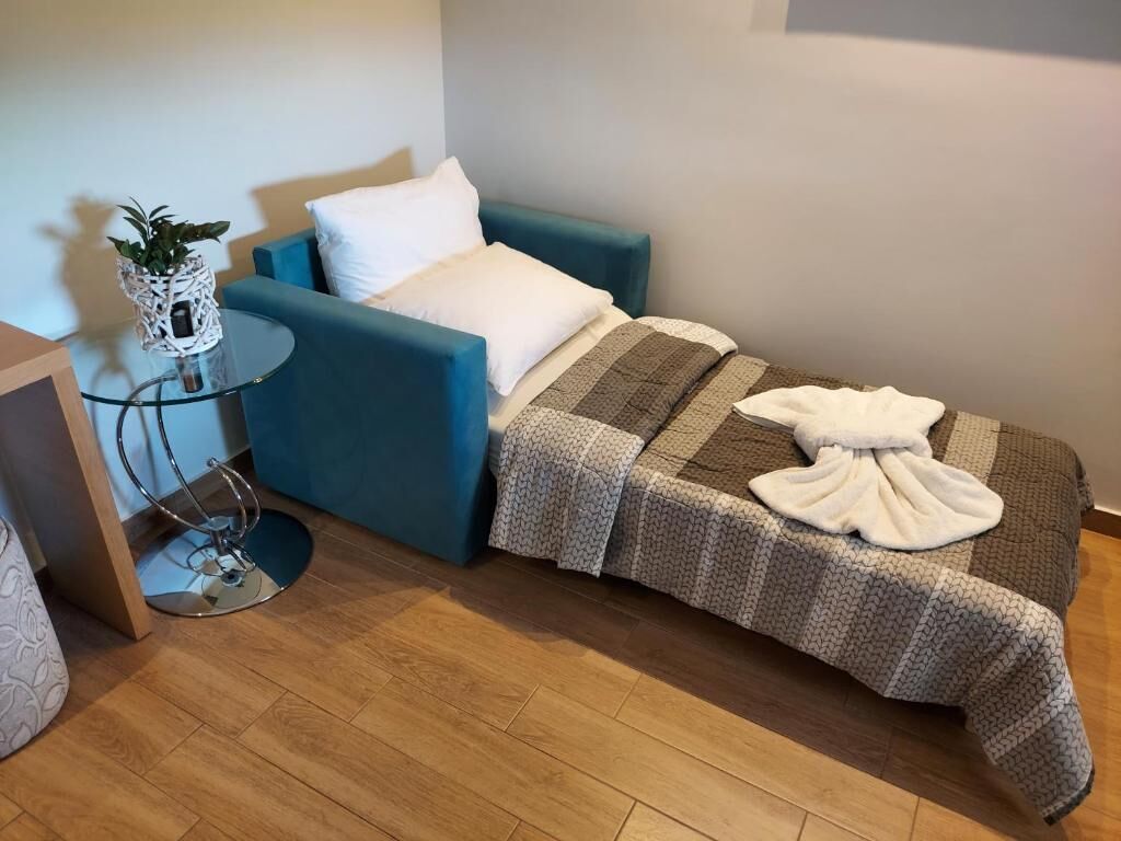 A small, cozy room with a single bed, a blue armchair, a glass side table with a plant, and a white pillow.