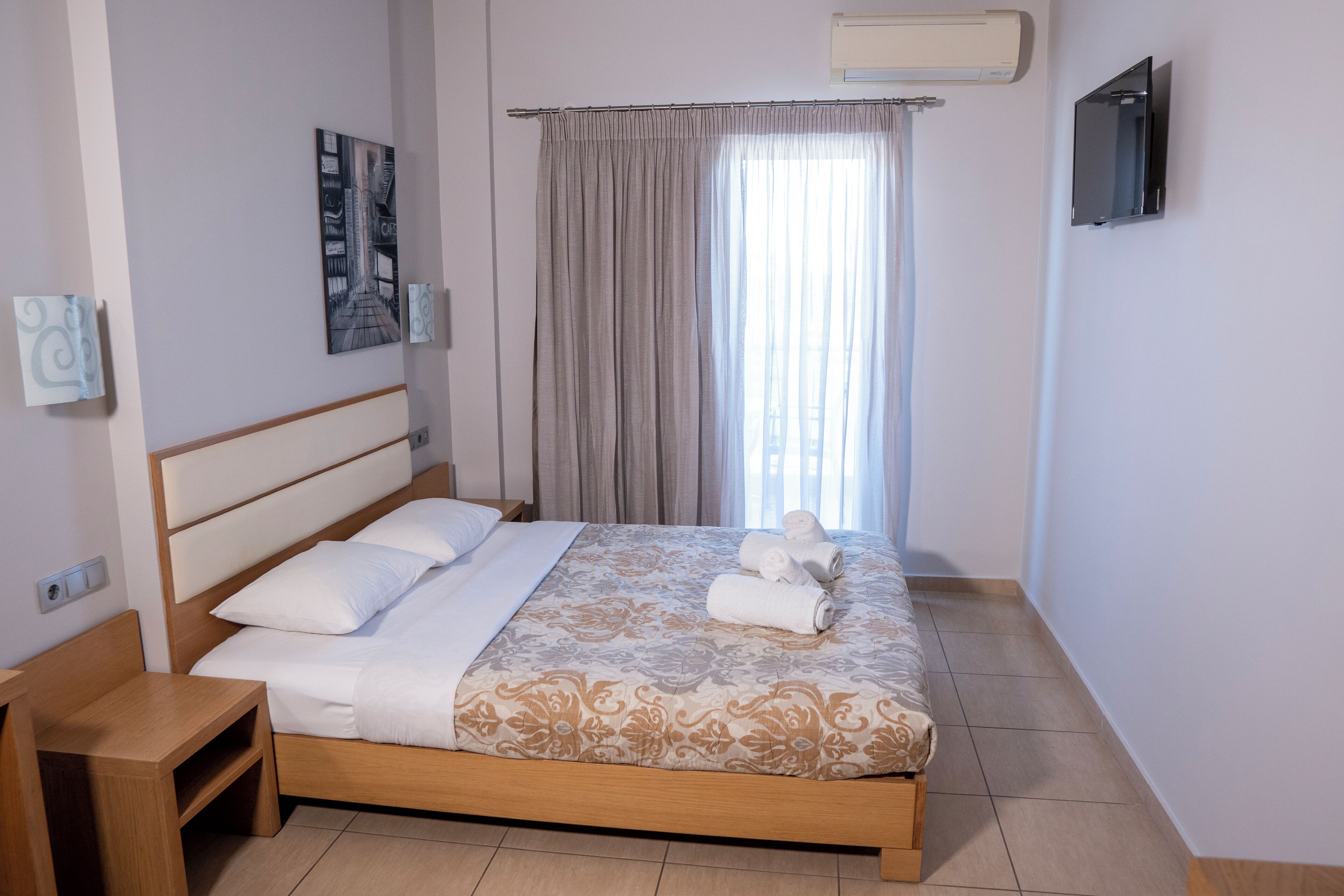 Una cama bien hecha con una colcha estampada, una mesita de noche, un televisor montado en la pared y una ventana con cortinas.