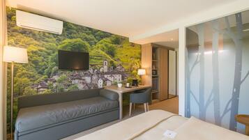Une chambre d’hôtel comprenant un lit, un canapé, un bureau avec une chaise, un téléviseur fixé au mur et une fresque représentant un village sur le mur.