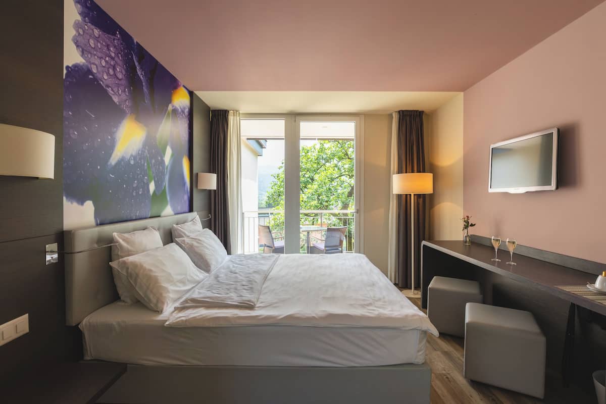 Una habitación de hotel con cama, escritorio, televisor y balcón con mobiliario exterior.