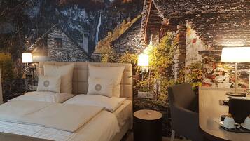 Un dormitorio con una cama grande, dos lámparas de noche, una mesita redonda con una silla y un mural en la pared que muestra un paisaje montañoso.
