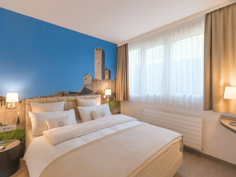 A hotel room with a large bed, two bedside lamps, a window with curtains, and a mural of a castle on the wall.