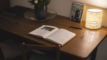 Een houten tafel met een open boek, een pen, een bloempot, een ingelijste foto en een brandende lamp.