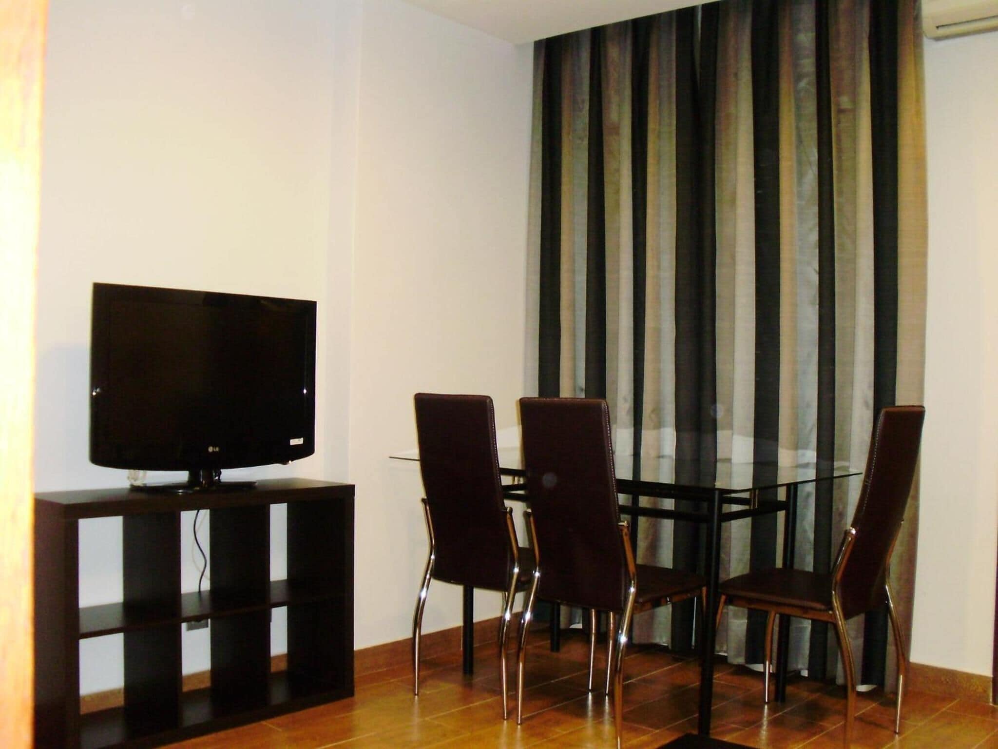 A modern living room with a flat-screen TV on a stand, a glass dining table with four chairs, and a large window with vertical blinds.