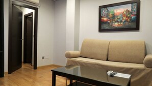 Een woonkamer met een beige bank, een glazen salontafel en een schilderij van een stadsgezicht aan de muur.