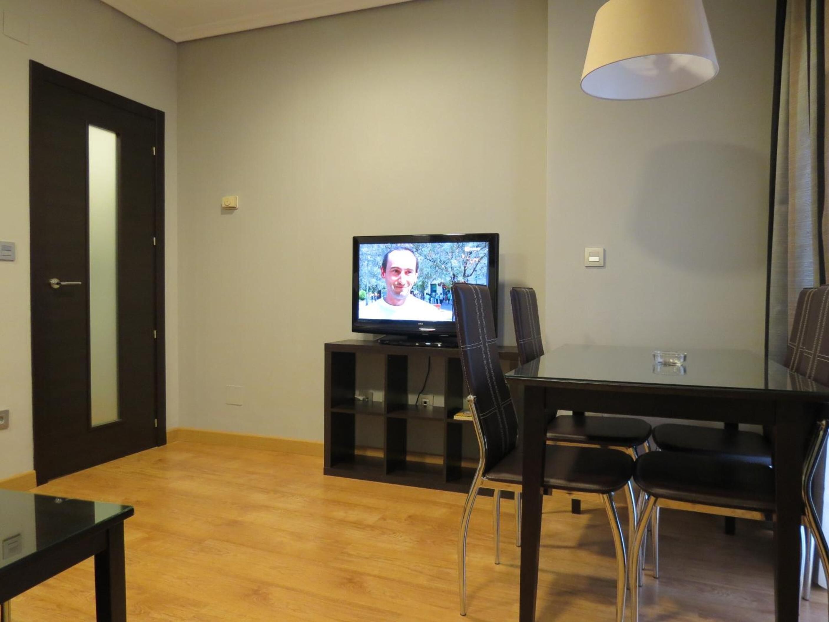 A modern living room with a flat-screen TV on a stand, a dining table with chairs, and a door.