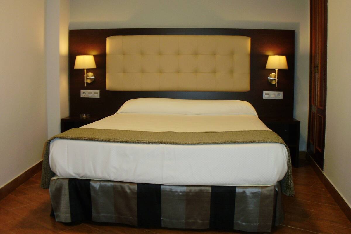Una habitación de hotel con una cama grande, dos lámparas de noche y un cabecero oscuro.