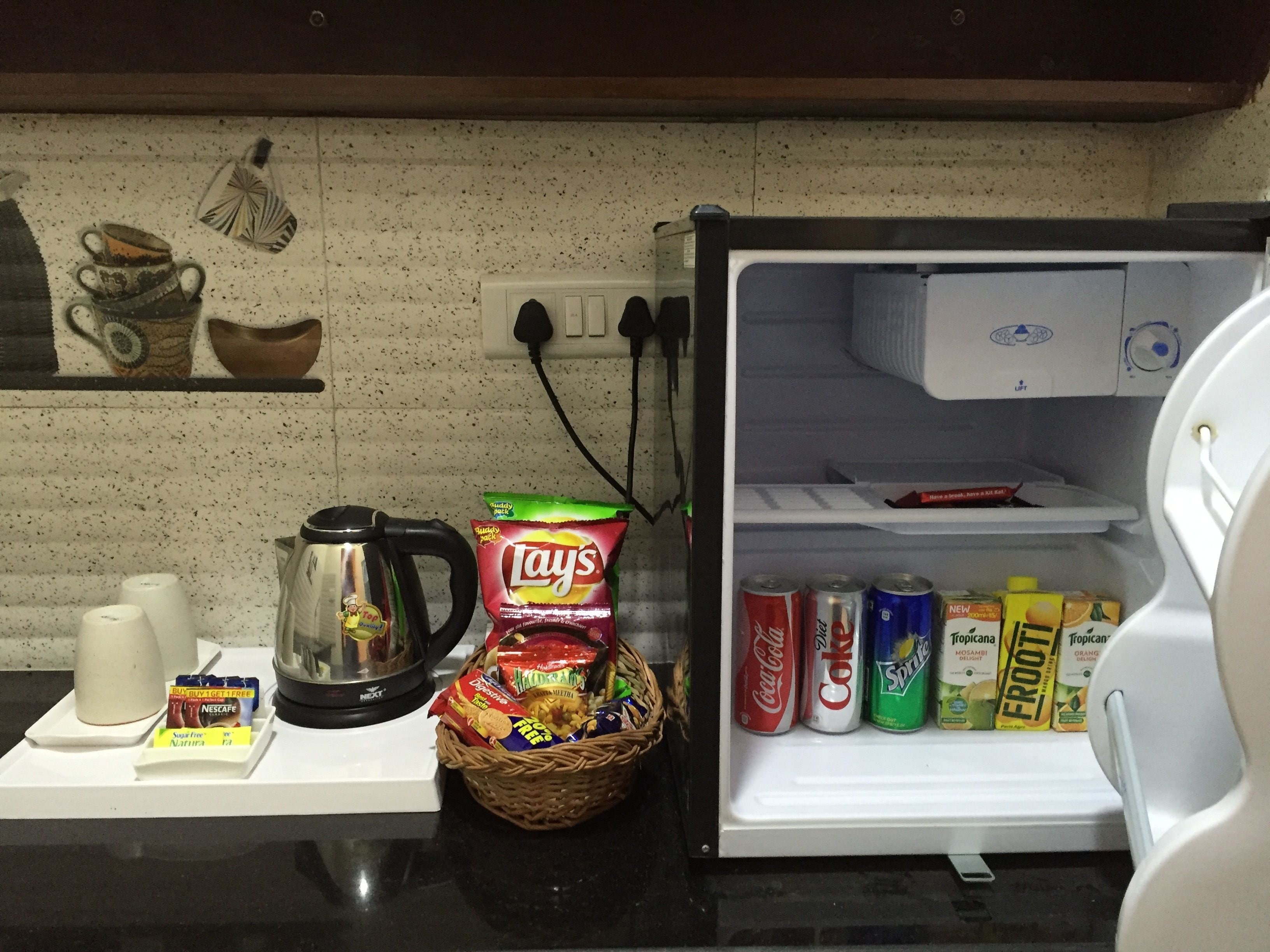 A kitchen counter with a coffee maker, a basket of snacks, and a refrigerator stocked with beverages.