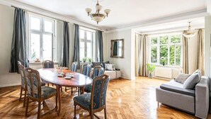 A spacious room with a wooden floor, a round dining table with chairs, a sofa, and a large window with curtains. - Apartments Happy Hours (Sopot)