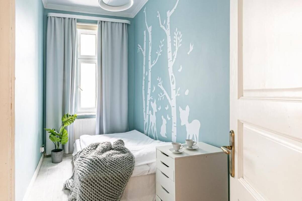 A bedroom with a bed, a dresser, a window with curtains, a potted plant, and a wall decal of trees and animals.