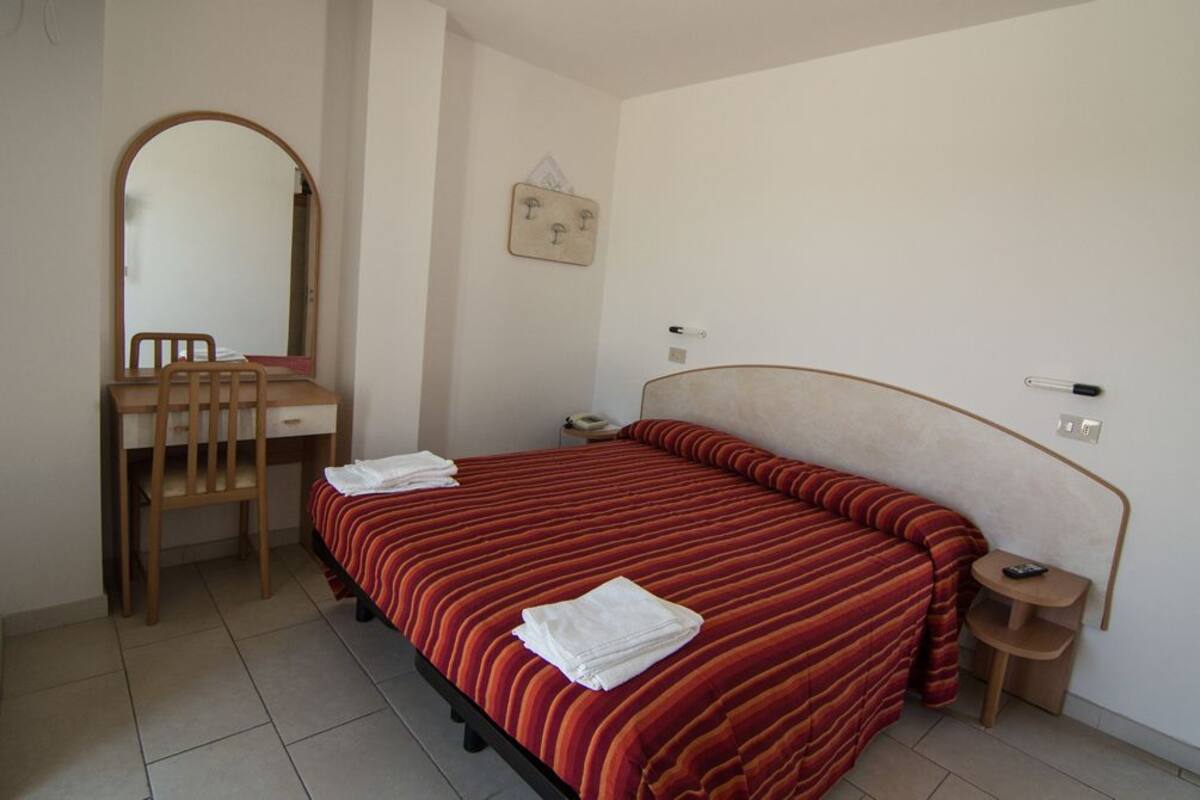 A bedroom with a bed, a desk, a chair, a mirror, and a small bedside table.
