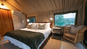 A bedroom with a bed, bedside table, lamp, and a wicker chair. - Sausage Tree Safari Camp (Hoedspruit)