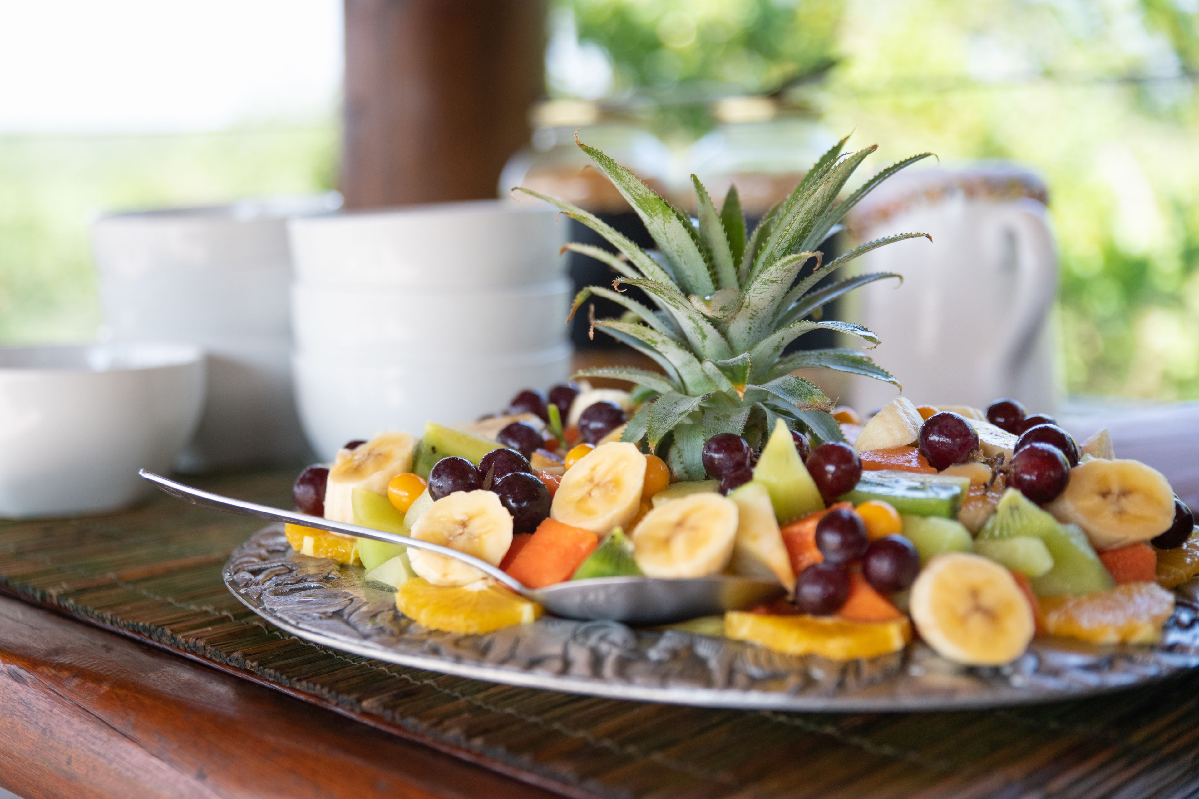 A fruit platter on a plate with a spoon, surrounded by white cups and saucers on a wooden table.