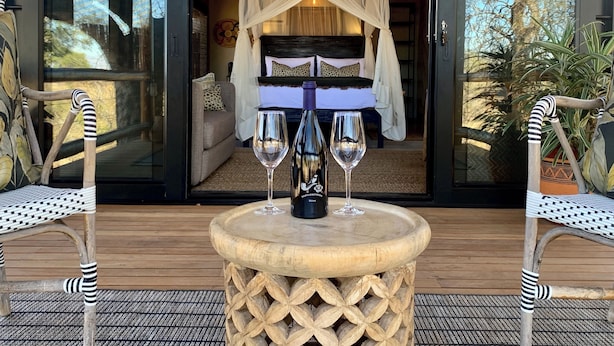 A room with a bed under a canopy, two chairs, a table with two wine glasses and a bottle, and a view of the outdoors. - Sausage Tree Safari Camp (Hoedspruit)