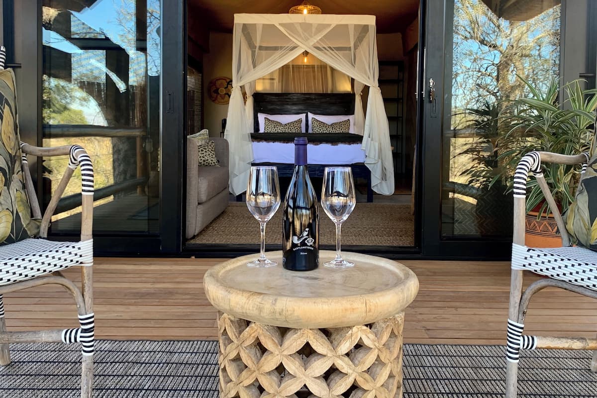 A room with a bed under a canopy, two chairs, a table with two wine glasses and a bottle, and a view of the outdoors.