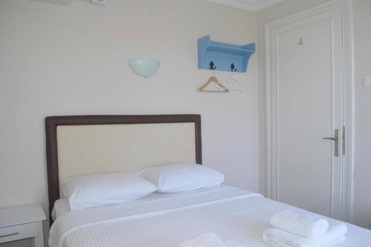 Un lit bien fait, avec du linge de lit blanc et des oreillers, une petite étagère fixée au mur sur laquelle repose un bol, et une porte sur la droite.